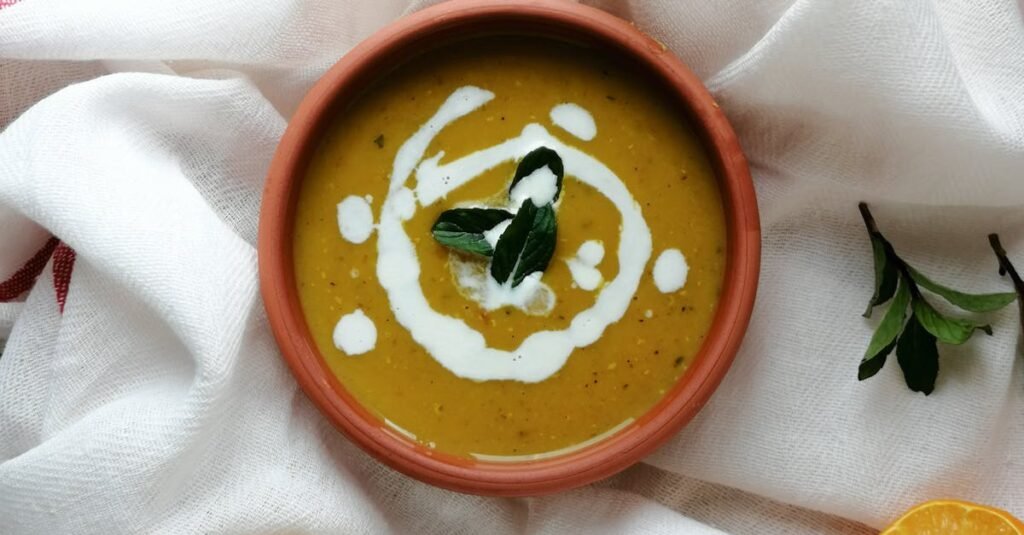 A close-up of creamy lentil soup with garnish, lemon slices, and bread on a white cloth.