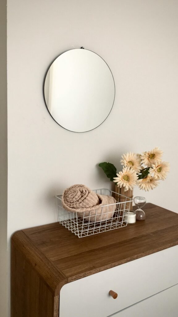 A minimalist home decor with a round mirror, daisies, and a wire basket on a wooden dresser.