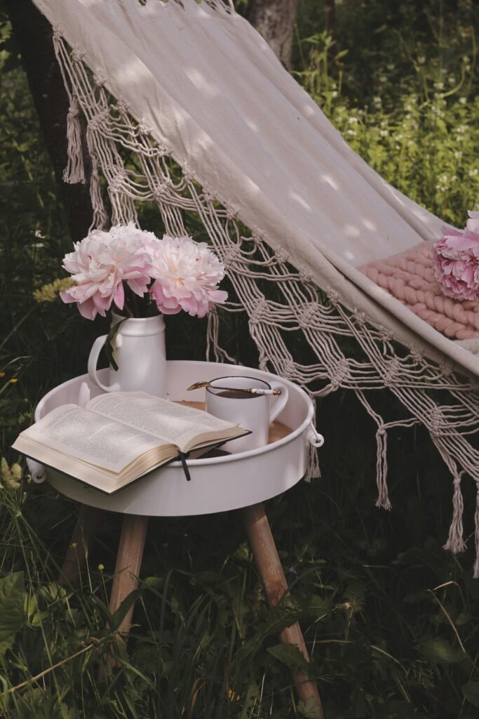 Serene outdoor setup with a hammock, pink flowers, an open book, and a cup on a tray table.
